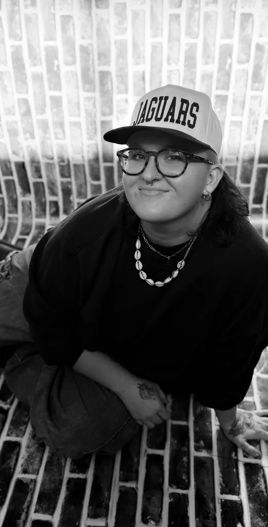 Black-and-white photo of Paige sitting on a painted brick floor in a Jaguars cap, round glasses and a shell necklace, smiling up at the camera.